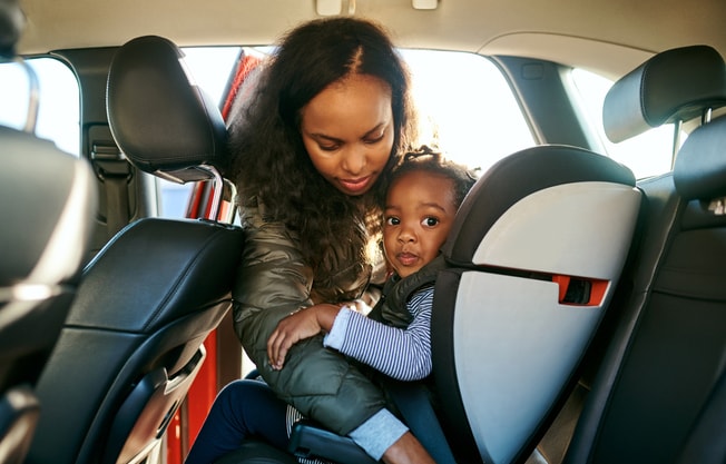 An African-Amerian mother buckles her daughter into a car seat as her daughter looks at the camera.