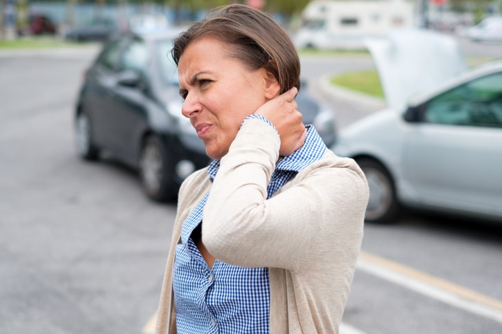 A woman is shown wincing in pain with her hand on her neck as two mangled cars are in the background.