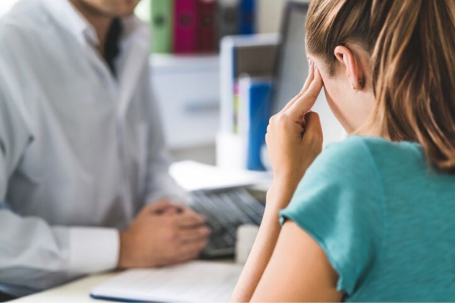 A woman sits with her hands to her temples with a doctor at his desk due to concussion symptoms.