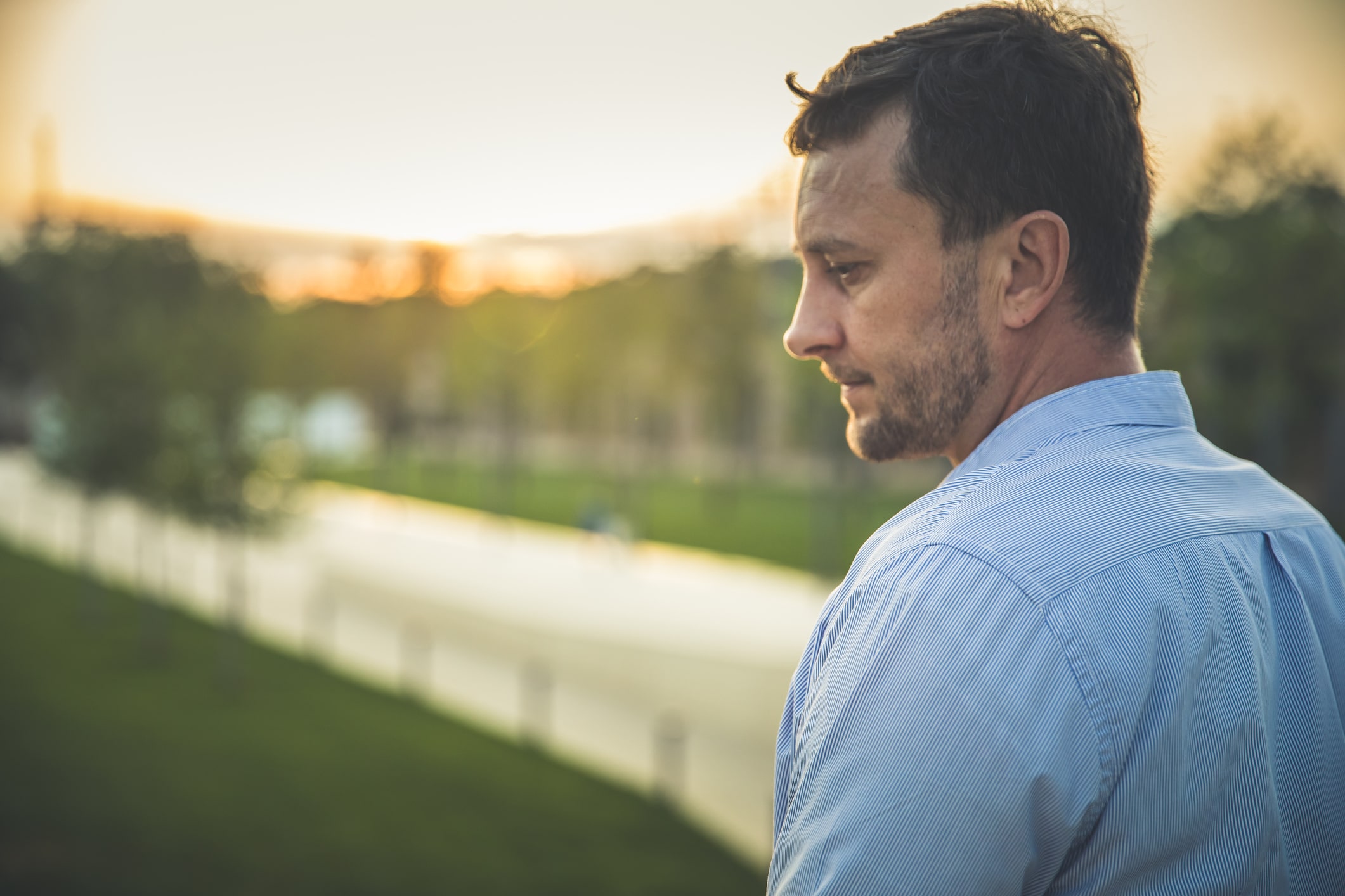 Man in blue collared shirt pondering near a pathway at sunset