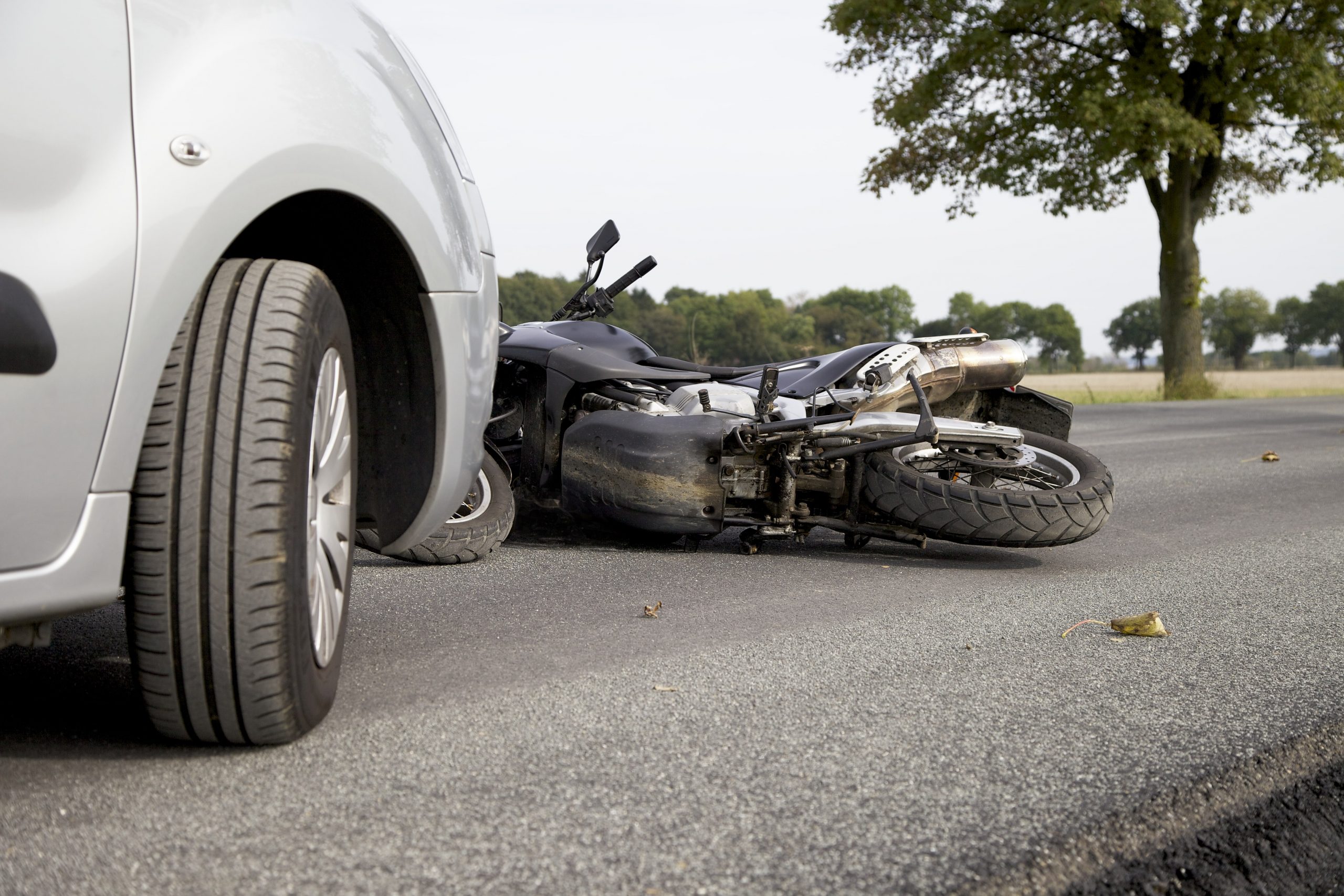 Silver car stopped in the road with a downed unmanned motorcycle in front of it after a crash