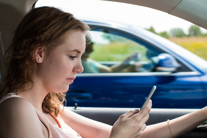 A young woman drives as she looks down at her phone. The driver next to her is not distracted.