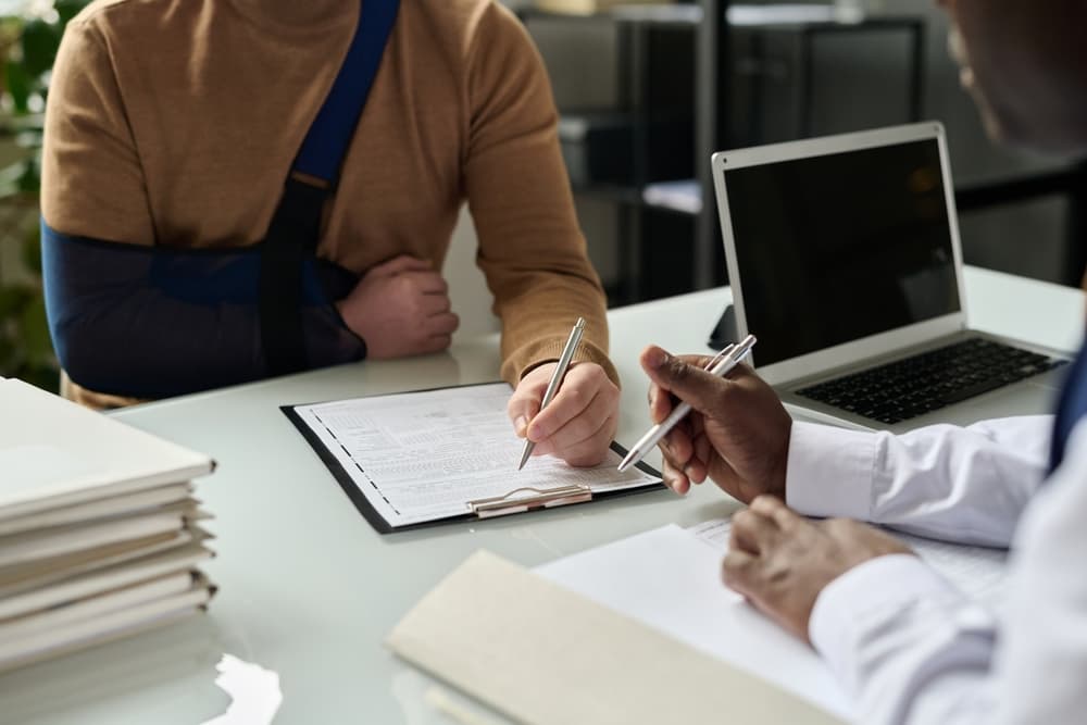 Close-up of a man completing a medical insurance form, with his injured arm in a sling visible in the background.