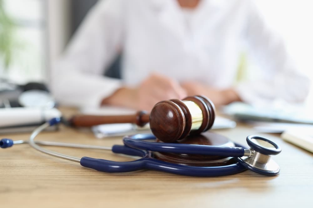Closeup of a gavel with a stethoscope; doctor in the background takes notes. Symbolizing the legal dimensions of healthcare and medical insurance payments.