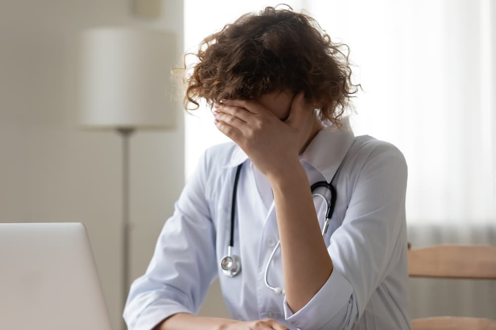 Close-up of distressed young woman doctor in white uniform covering face. Expresses stress, guilt, and despair in a hospital setting.