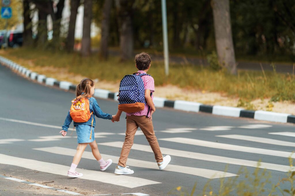 Two children with backpacks holding hands while safely crossing the street at a pedestrian crosswalk.