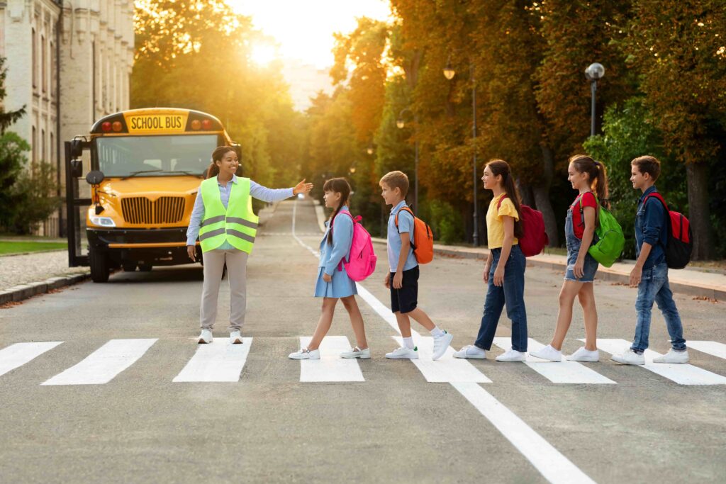 School crossing guard helping children with backpacks safely cross the street in front of a school bus.
