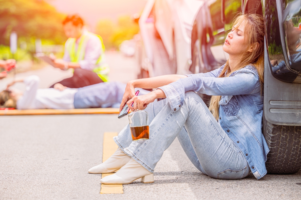 A woman sitting injured on the road after a crash while holding a liquor bottle, with paramedics assisting another victim in the background, as seen in cases handled by a Glastonbury drunk driving accident lawyer.